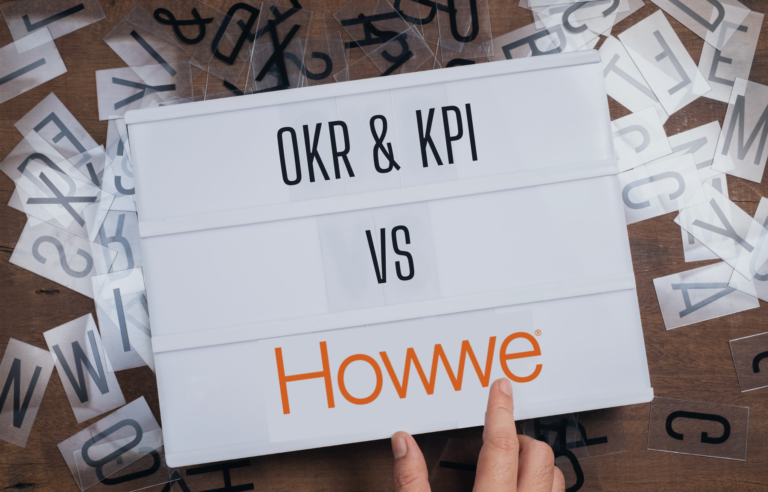 A signboard displaying "OKR & KPI VS" in bold black letters on the top two rows, with the Howwe® logo in orange at the bottom. The signboard is surrounded by scattered transparent letter sheets on a wooden surface, and a hand is pointing at the Howwe® logo.